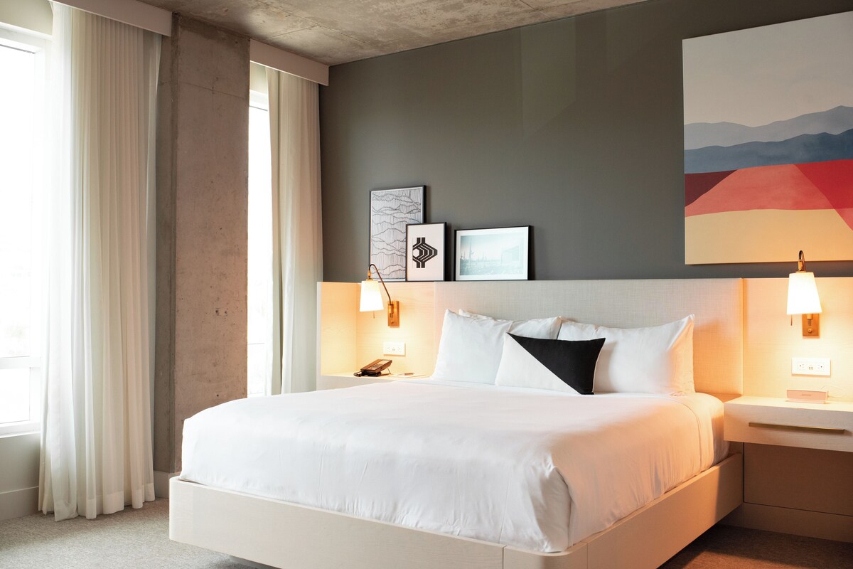 A modern hotel room with a large bed, bedside tables, a nightstand, and a window with curtains.
