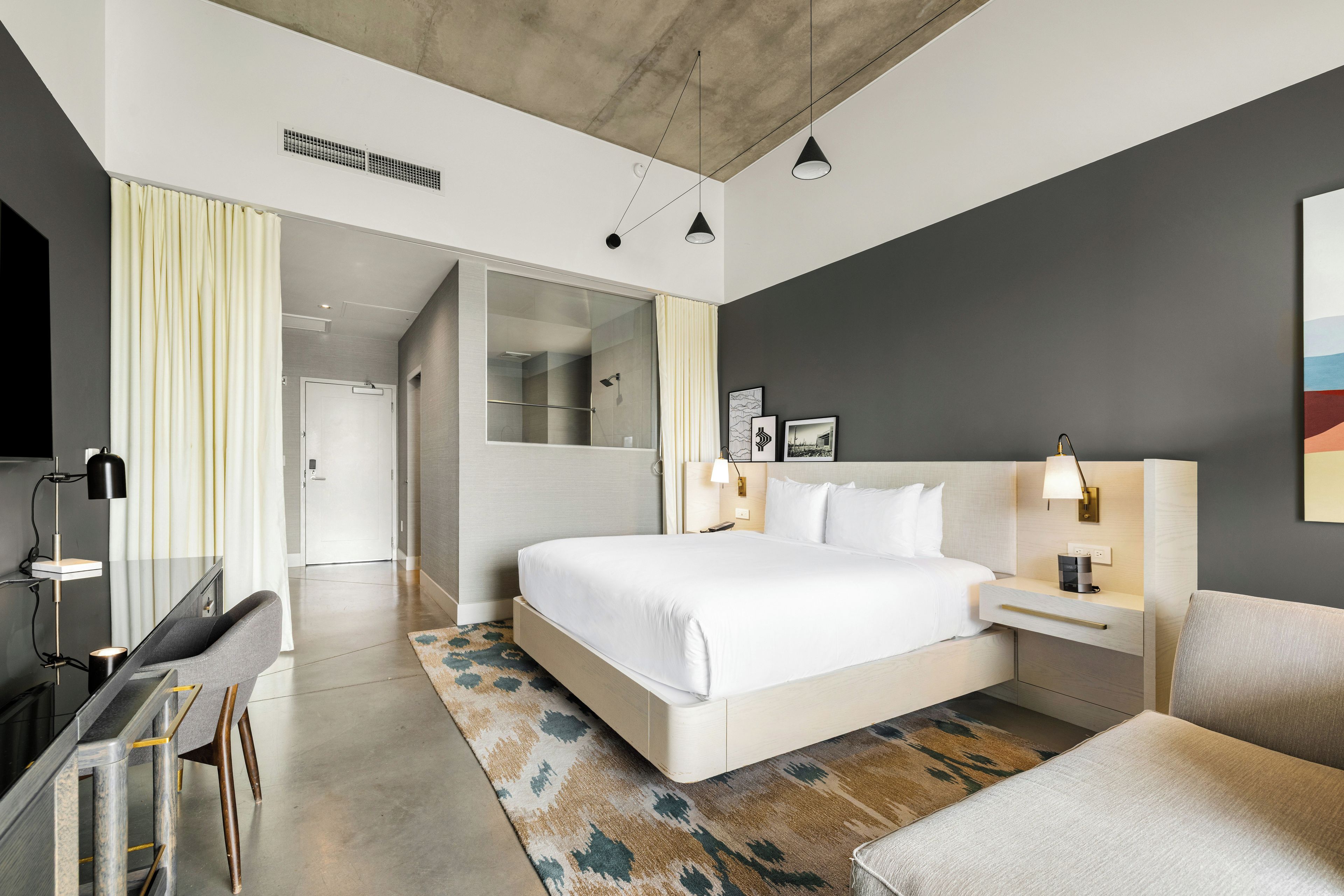 A modern hotel room with a large bed, bedside tables, a desk, a chair, a shower area, and a view of the hallway.