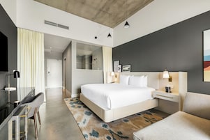 A modern hotel room with a large bed, bedside tables, a desk, a chair, a shower area, and a view of the hallway.
