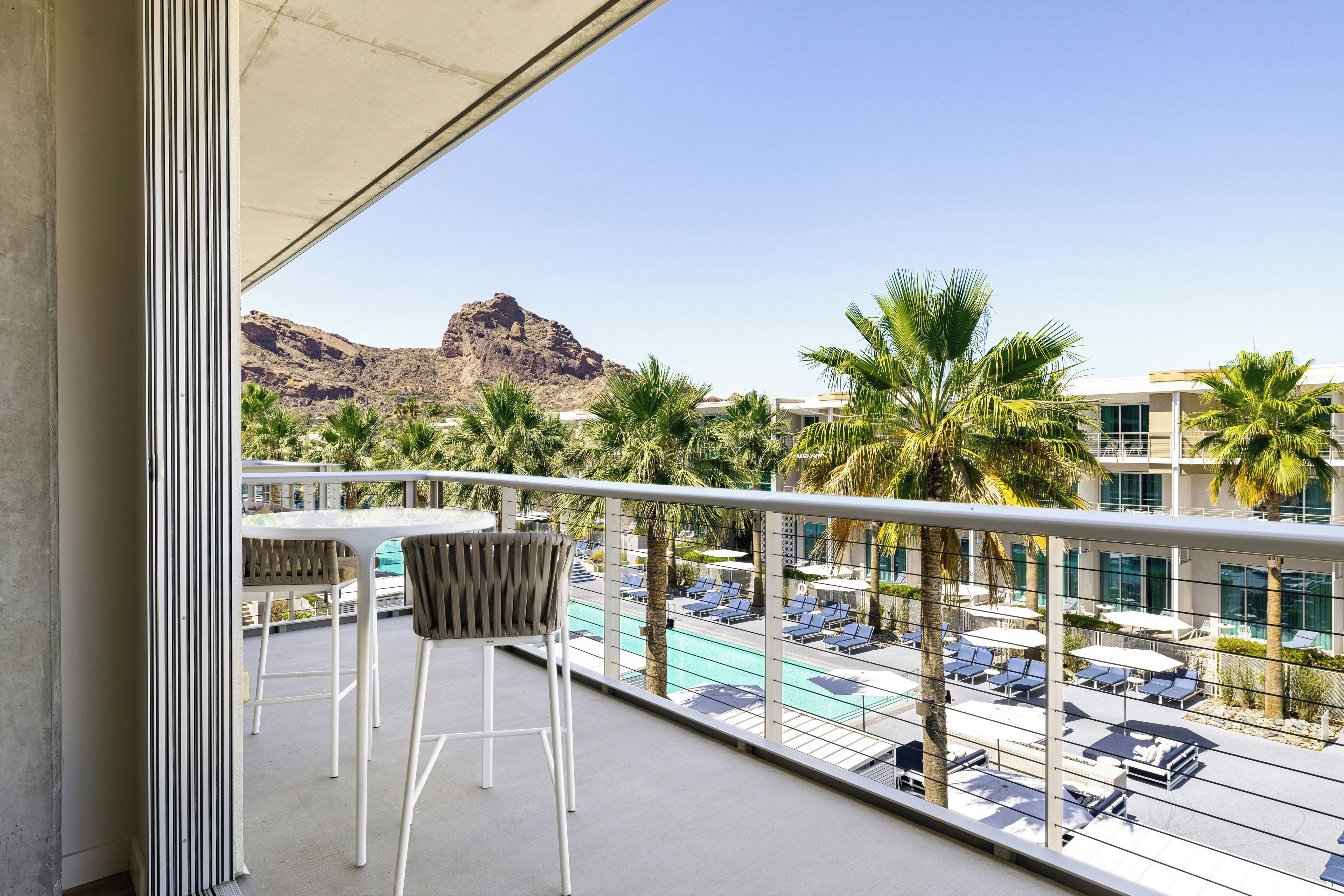 Un balcon avec deux chaises, un espace piscine avec des chaises longues et des parasols, et une vue sur les montagnes.
