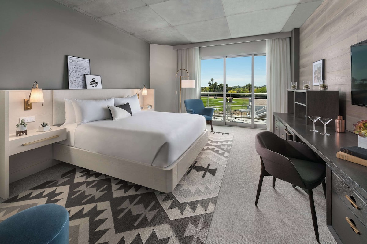 A modern hotel room with a large bed, a desk with a chair, a TV, and a balcony with a view.