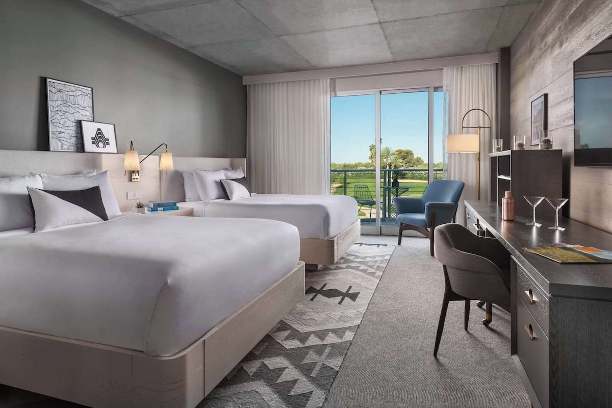 A hotel room with two beds, a desk with a chair, a TV, and a balcony with a view of the outdoors.