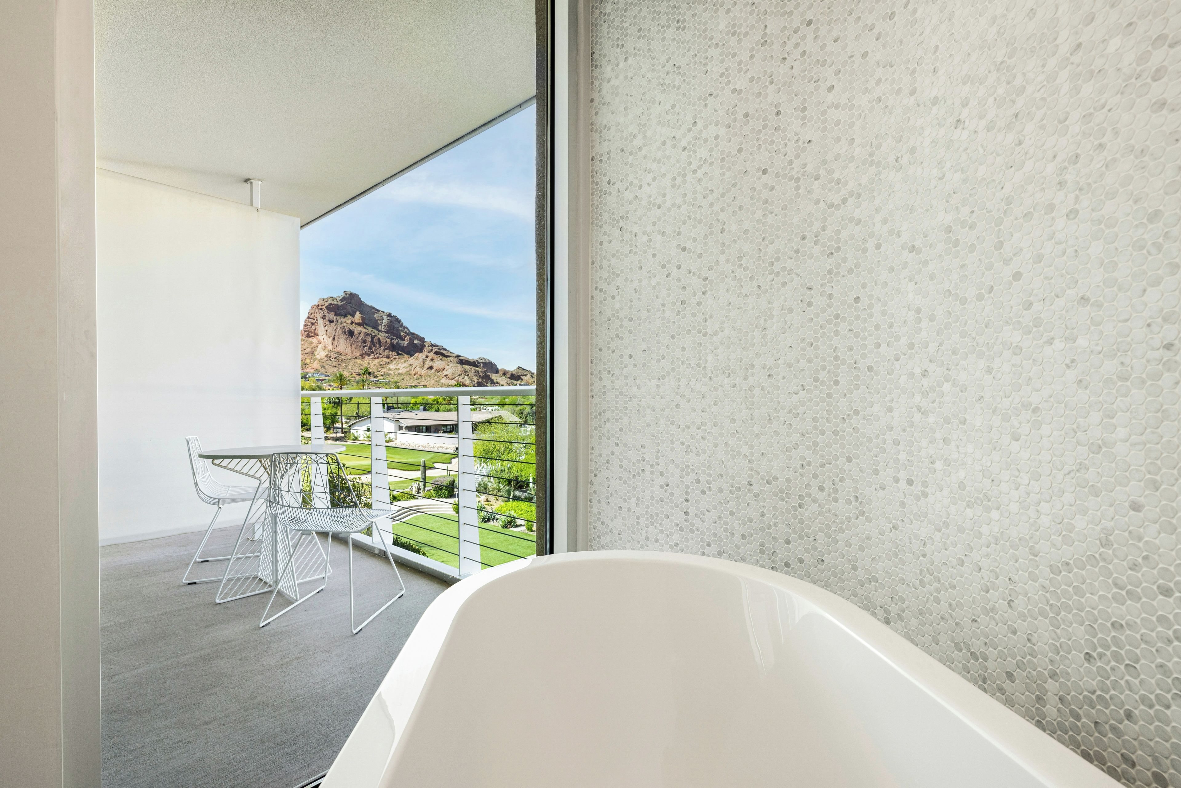 Une salle de bain moderne avec une baignoire sur pieds, une vue sur un paysage montagneux et un balcon agrémenté d’une chaise et d’une table en métal.