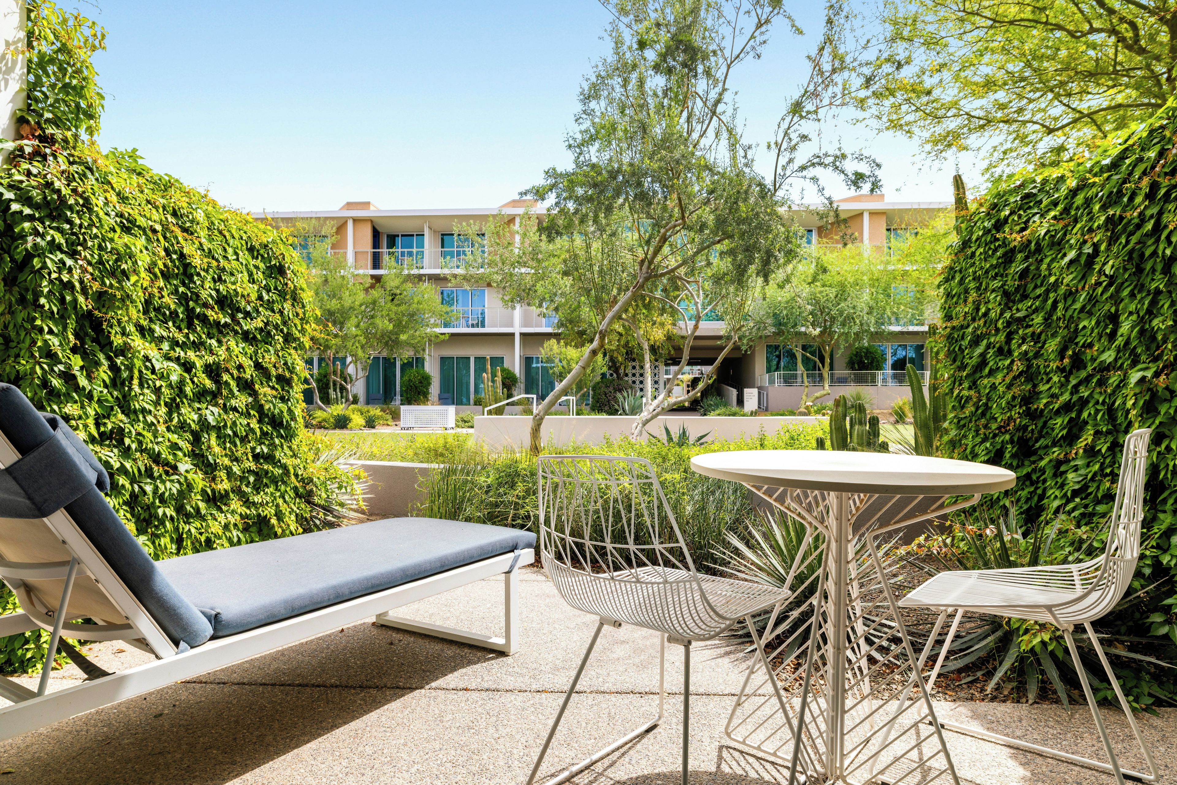 Een zonnig terras buiten met een lounge stoel, een tafel en stoelen, omgeven door weelderige beplanting en een gebouw op de achtergrond.