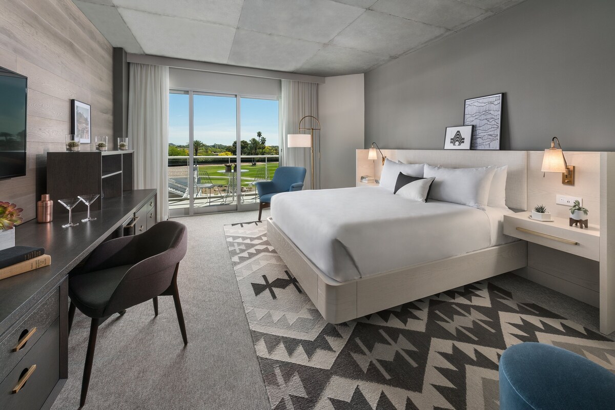 A modern hotel room with a large bed, a desk with a chair, a TV, and a balcony with a view of the outdoors.