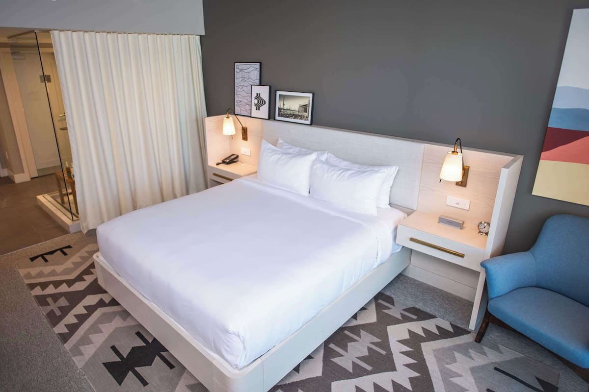 A hotel room with a large bed, two bedside tables, a nightstand, a blue armchair, and a patterned rug.