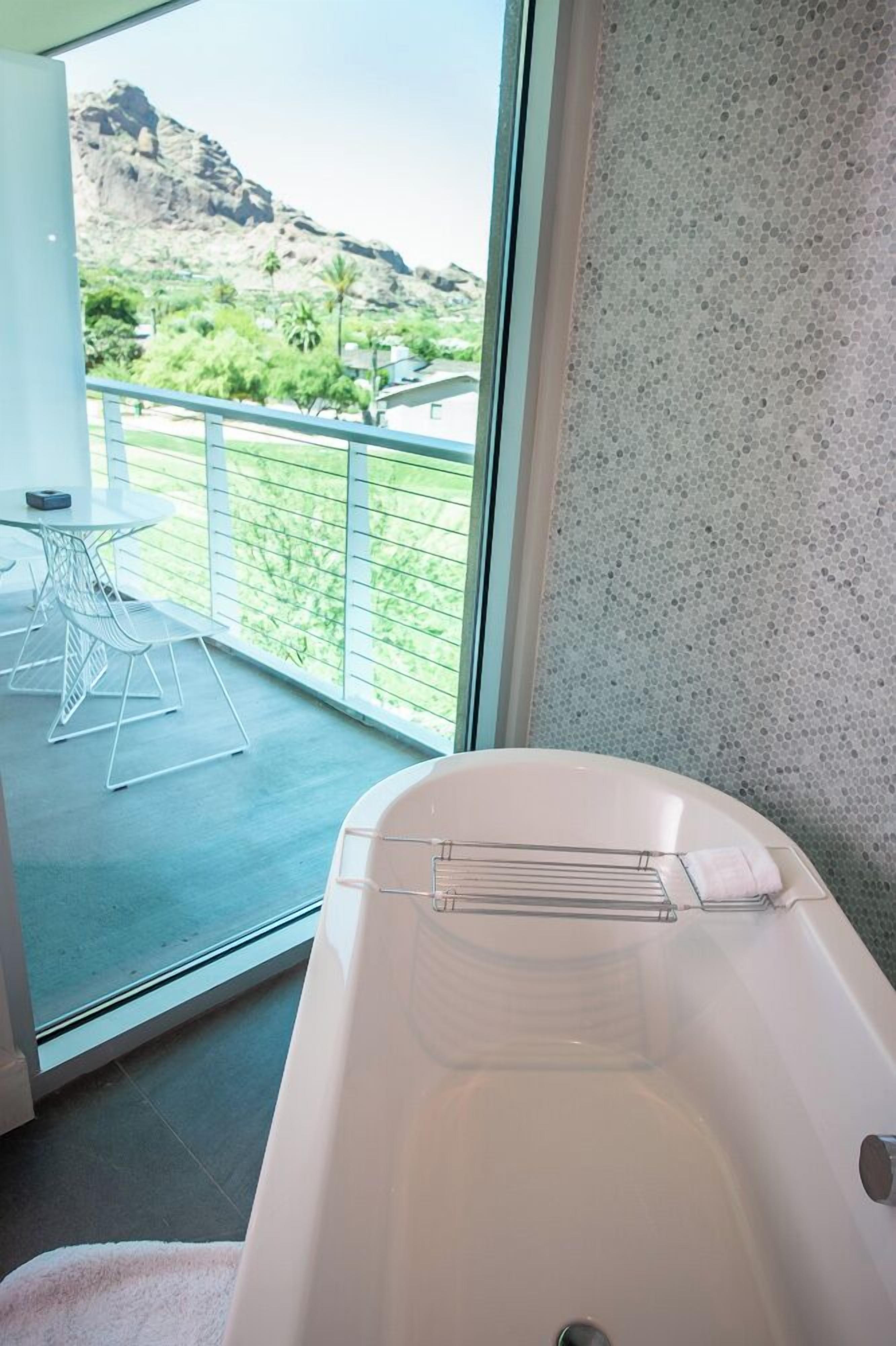 Un baño moderno con una bañera independiente, una mesita con sillas y un balcón con vistas a un paisaje montañoso.