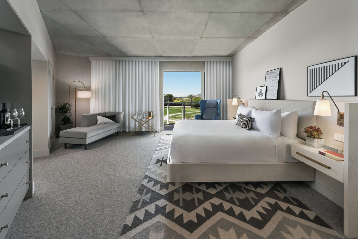 A modern hotel room with a large bed, a sofa, a desk with a lamp, and a view of a landscaped area.
