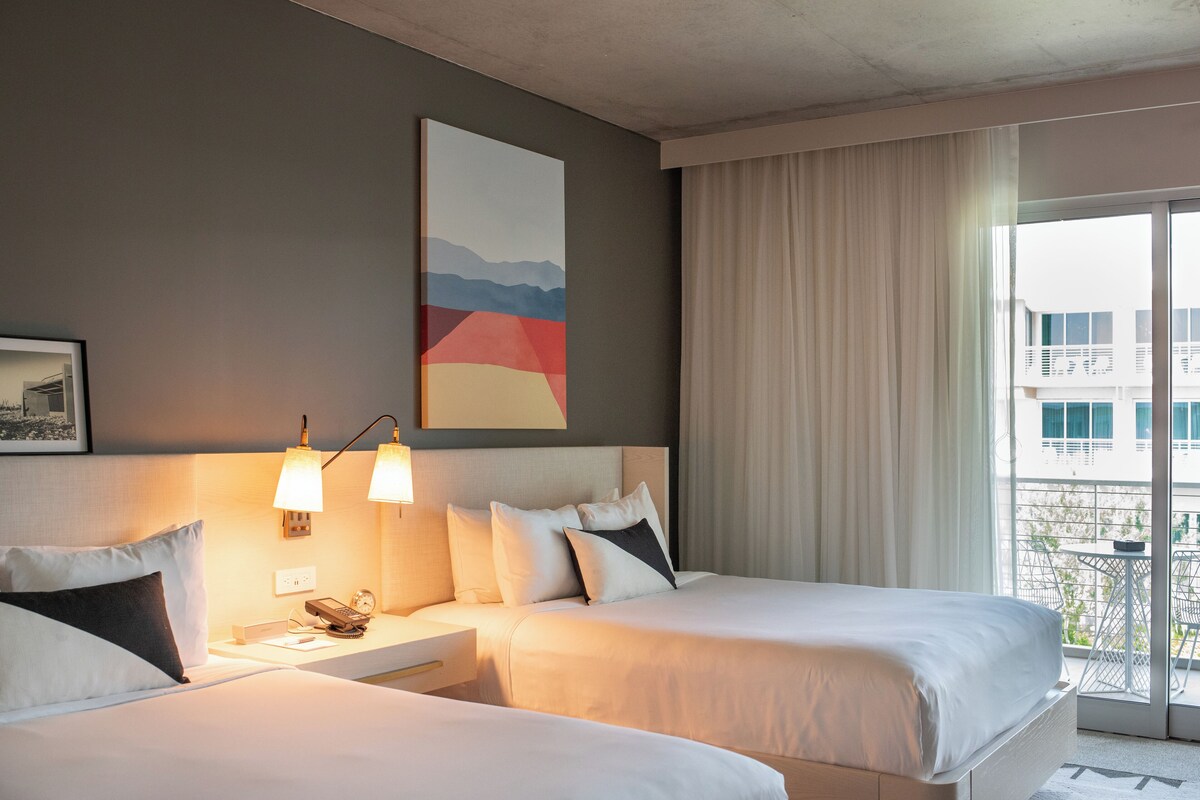 A hotel room with two beds, a nightstand with a lamp, a painting on the wall, and a view of the outside balcony.