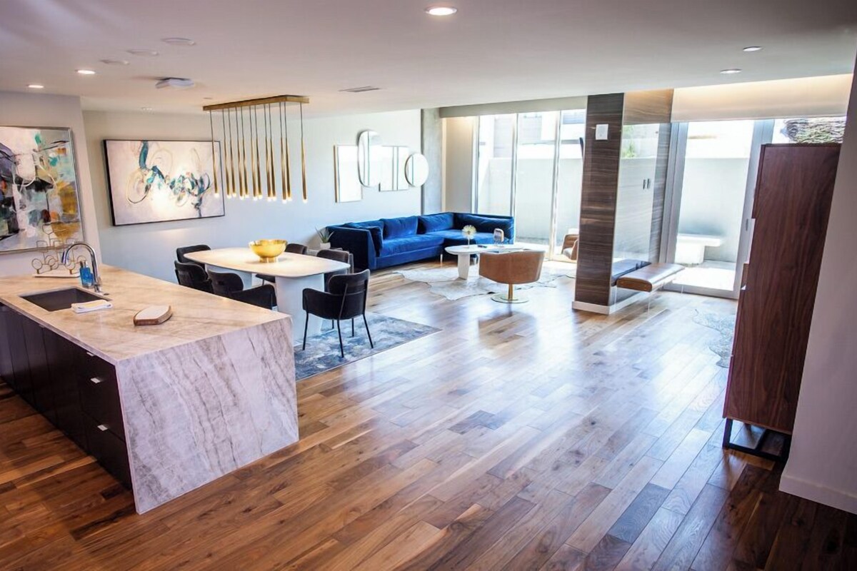 A modern kitchen with a marble island, a dining area with a round table, a blue sofa, and a large window providing natural light.