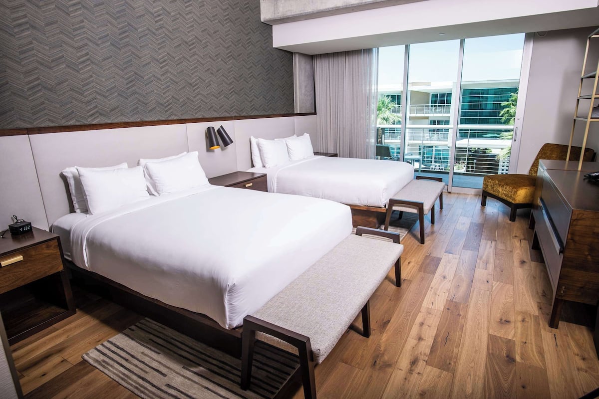A hotel room with two beds, a desk, a chair, and a view of a building through large windows.