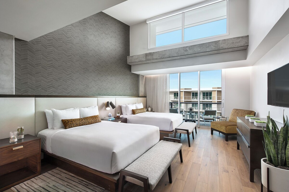 A modern hotel room with two beds, a seating area, a TV, and a balcony with a view.