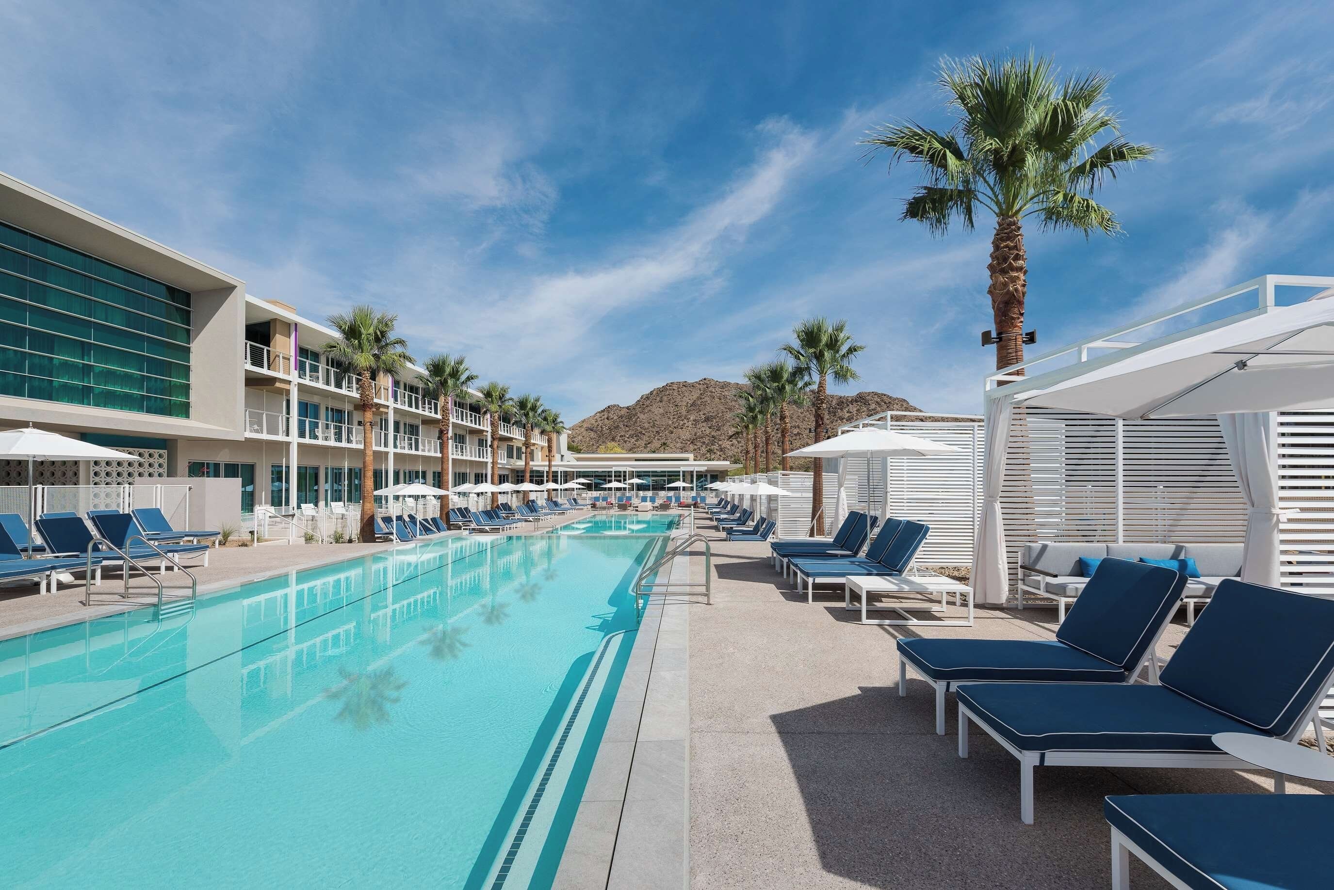 A pool area with lounge chairs and umbrellas, a building with balconies, and palm trees.