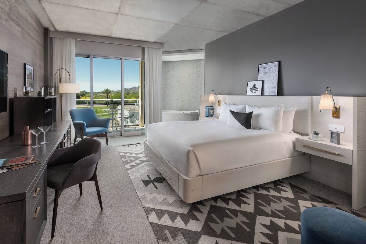 A modern hotel room with a large bed, a desk with a chair, a TV, and a balcony with a view.