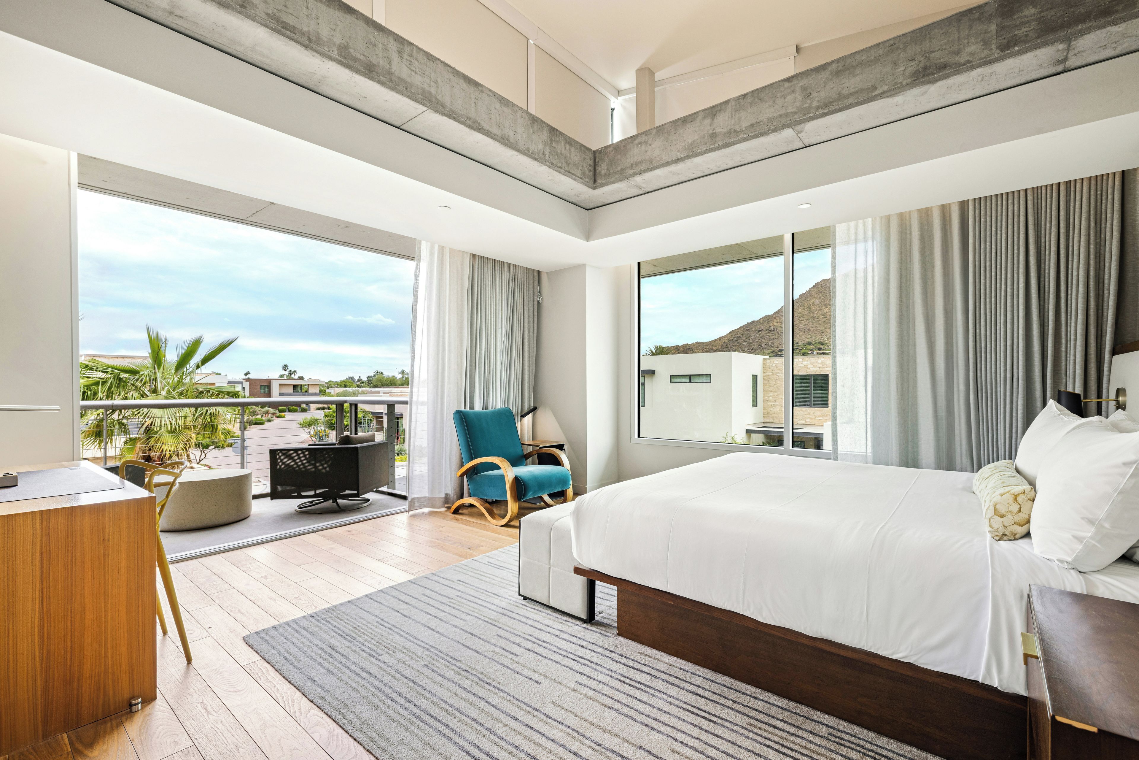 A modern bedroom with a large bed, a desk, and a chair, featuring a balcony with a view of the landscape.