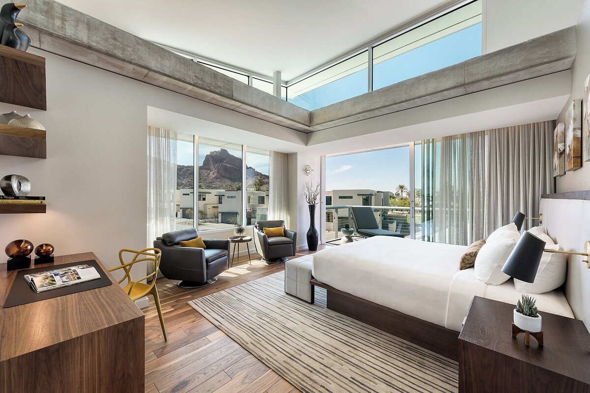 A modern hotel room with a large bed, a desk, a chair, and a balcony with a view of the mountains.