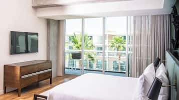 A modern hotel room with a large bed, a wooden dresser, a TV, and a balcony with a view of palm trees.