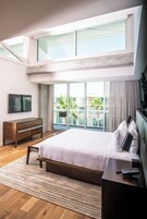 Una habitación de hotel moderna con una cama grande, un aparador de madera, un televisor y un balcón con vistas a palmeras.