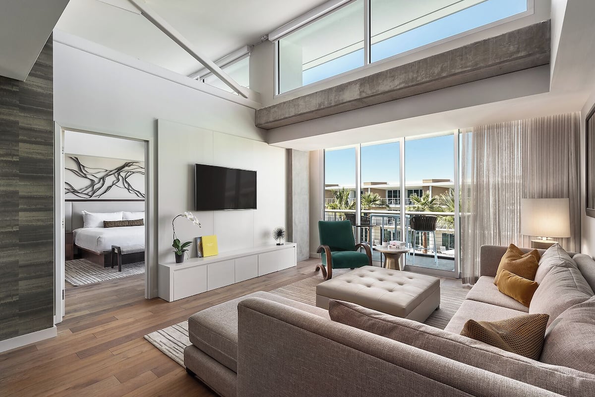 A modern hotel room with a large bed, a TV, a balcony with a view, and a seating area with a sofa and armchairs.