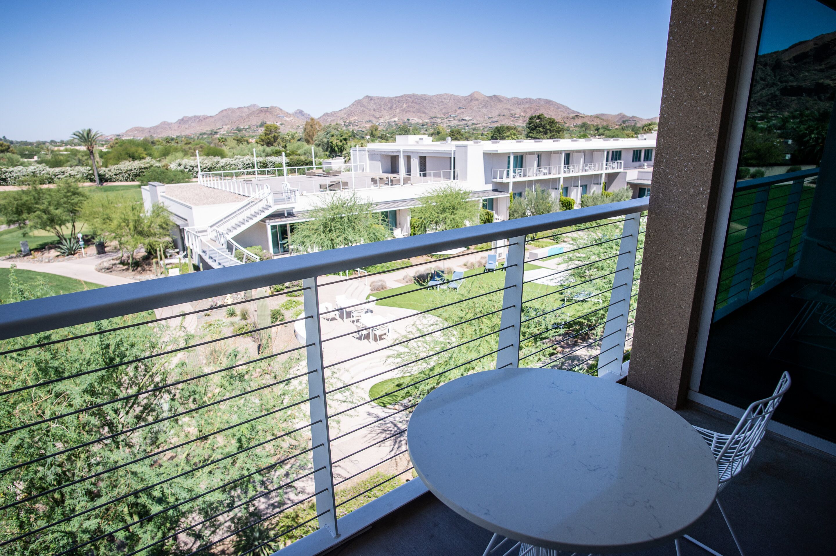 A balcony with a white table and chairs, overlooking a landscaped area with a pool and a building in the background.