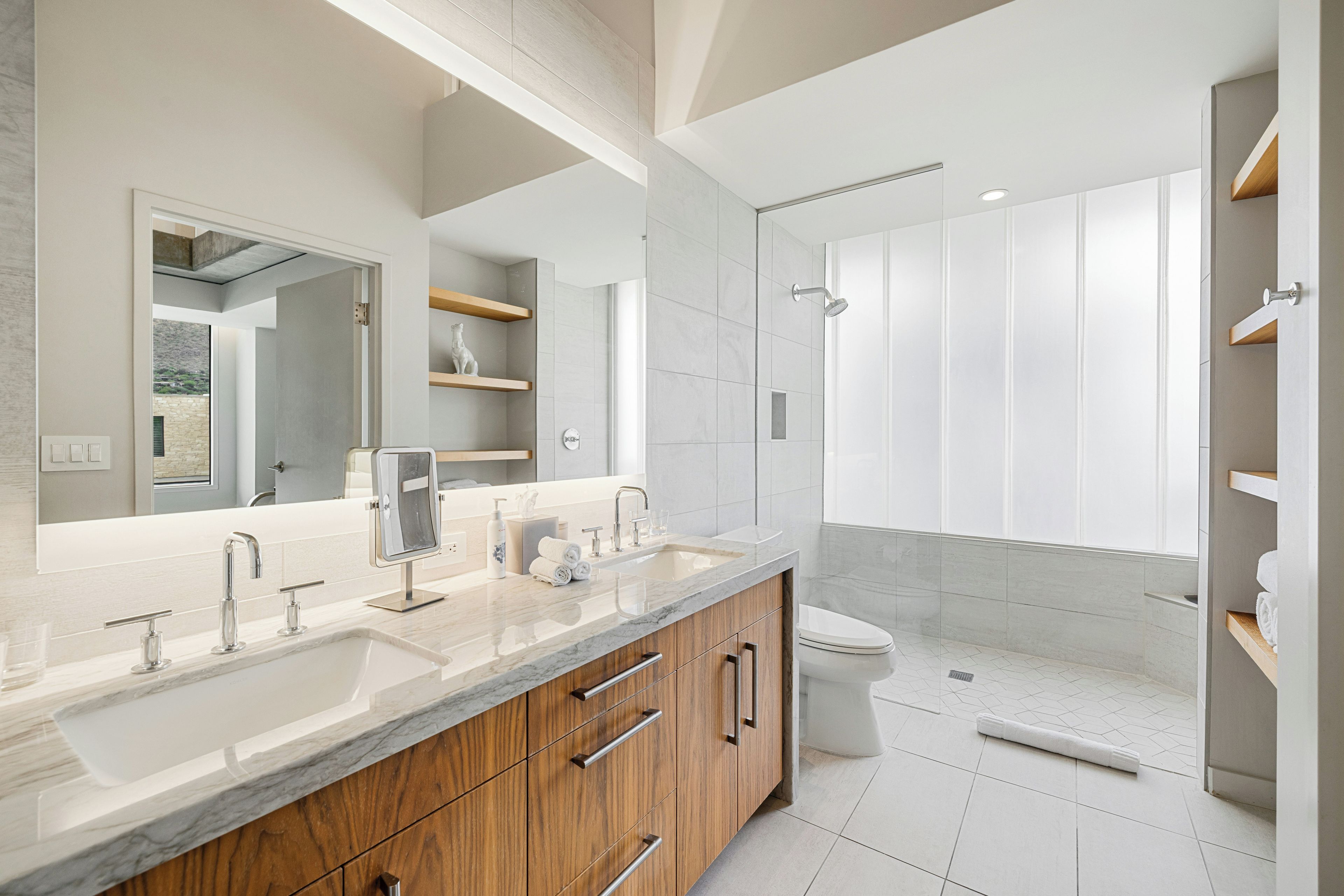 A modern bathroom with a large mirror, dual sinks, wooden cabinetry, and a walk-in shower.