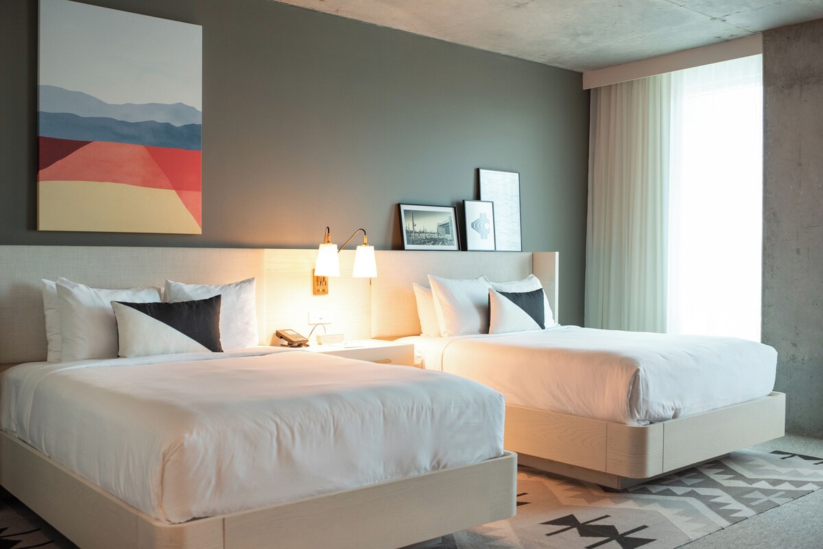 A hotel room with two beds, a nightstand with a lamp, a wall-mounted phone, and framed pictures.