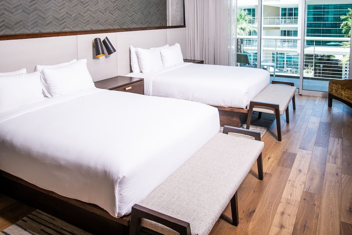 A modern hotel room with two beds, a large window, and a view of the city.