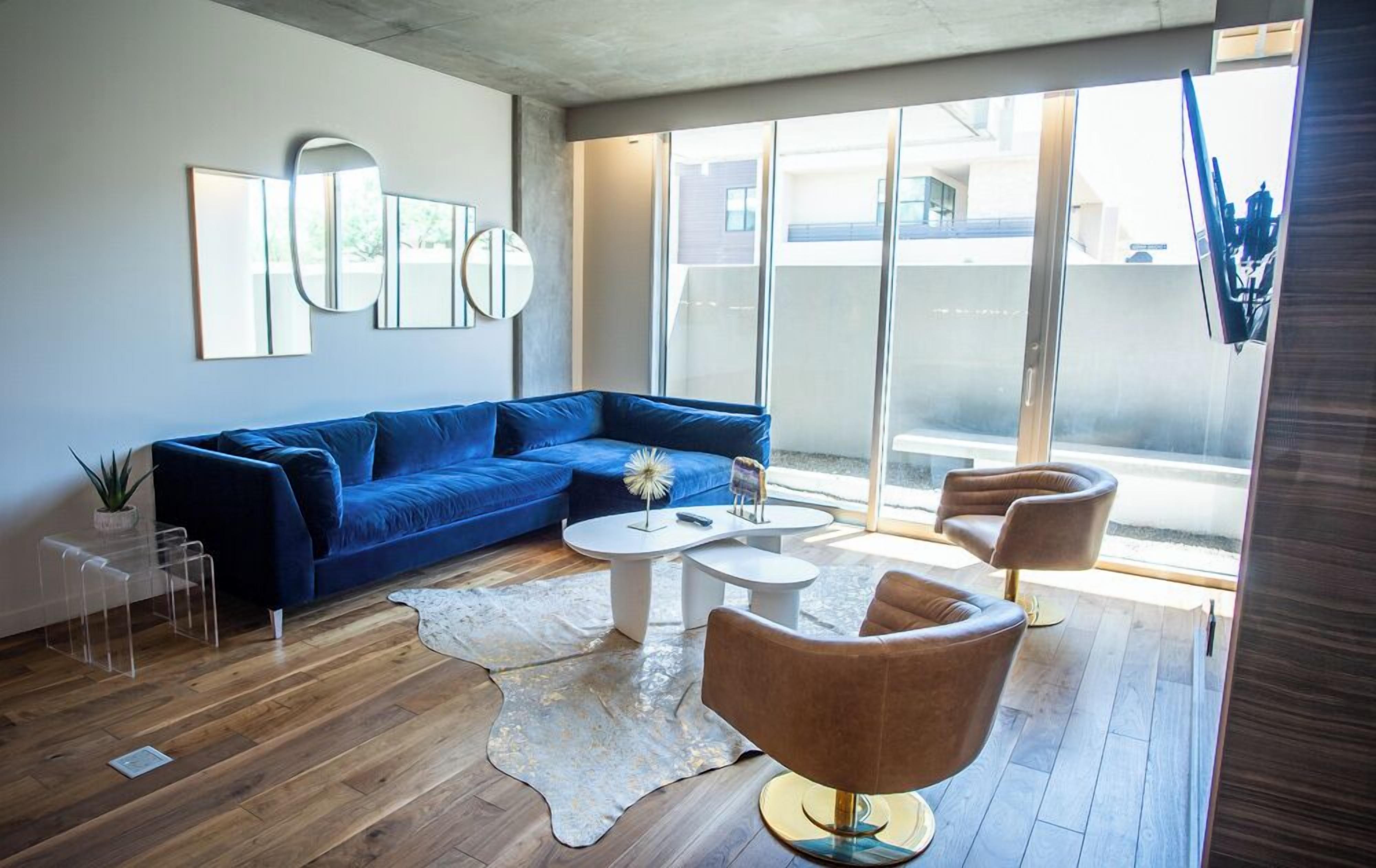 Una sala de estar moderna con un sofá azul, dos mesas de centro blancas y un sillón marrón. Hay un ventanal con vistas a edificios.