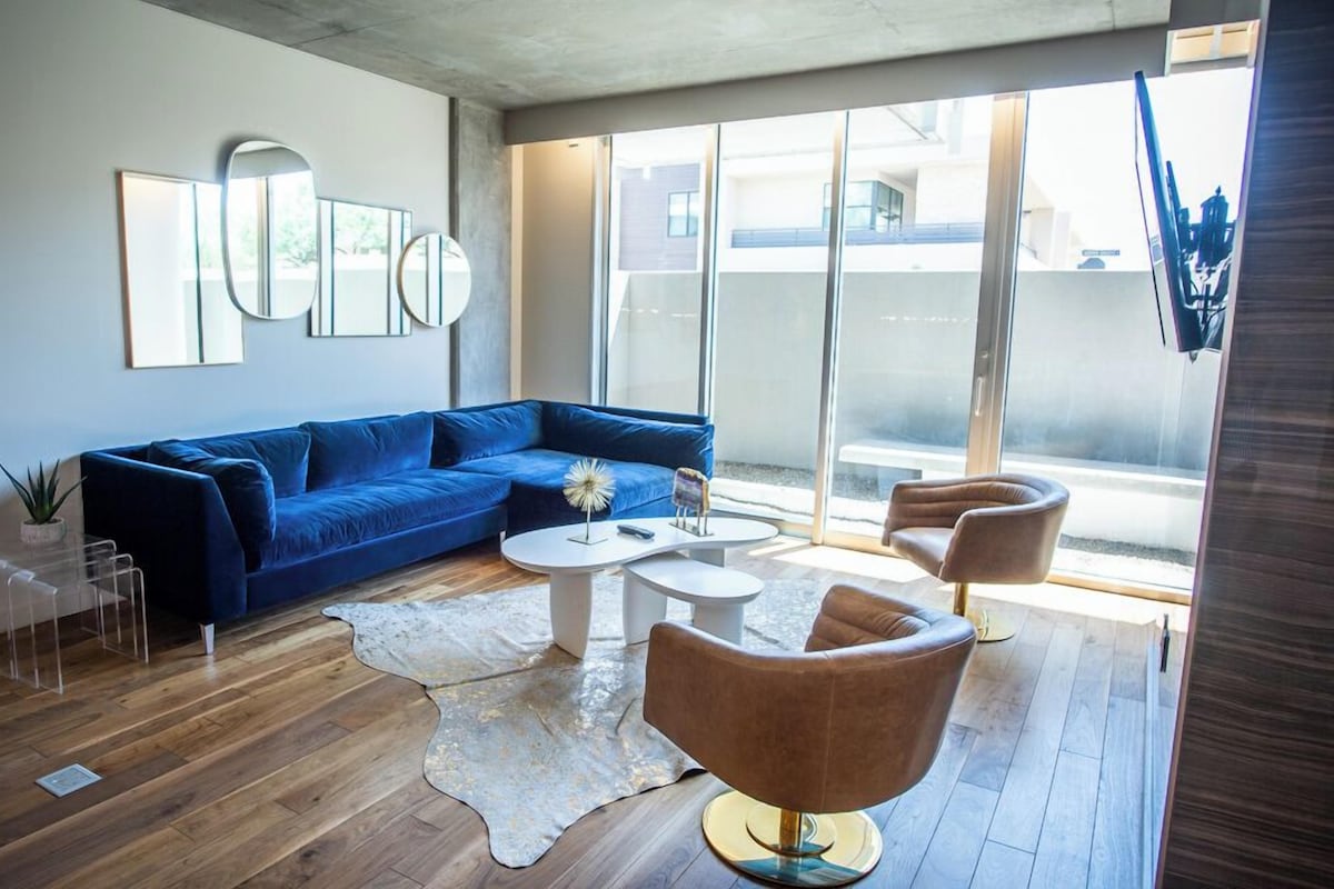 A modern living room with a blue sofa, two white coffee tables, and a brown armchair. There is a large window with a view of buildings outside.