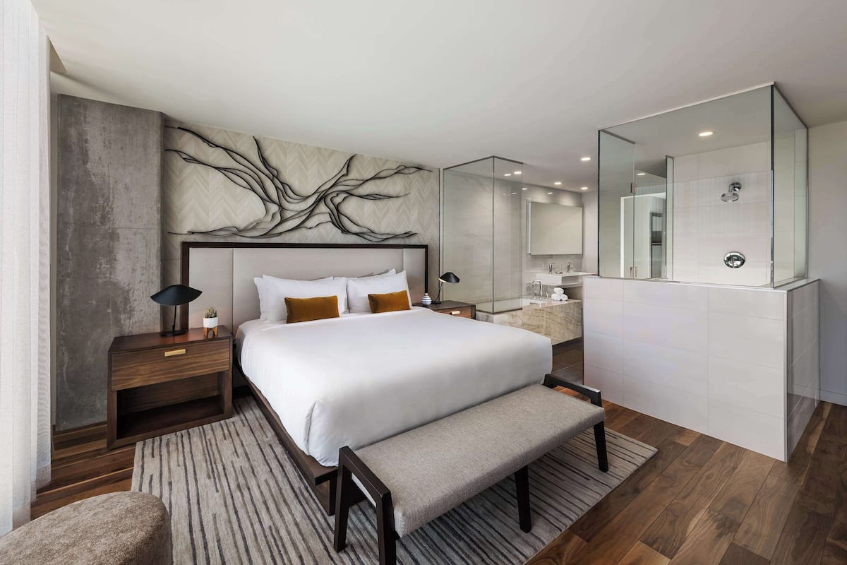 A modern bedroom with a large bed, a bench, a nightstand, a wall-mounted artwork, and a bathroom with a large mirror and sink.