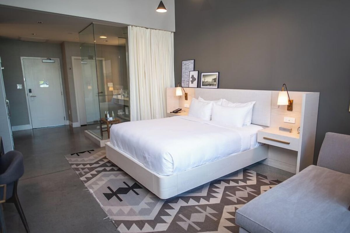 A modern hotel room with a large bed, bedside tables, a chair, and a glass-enclosed bathroom.