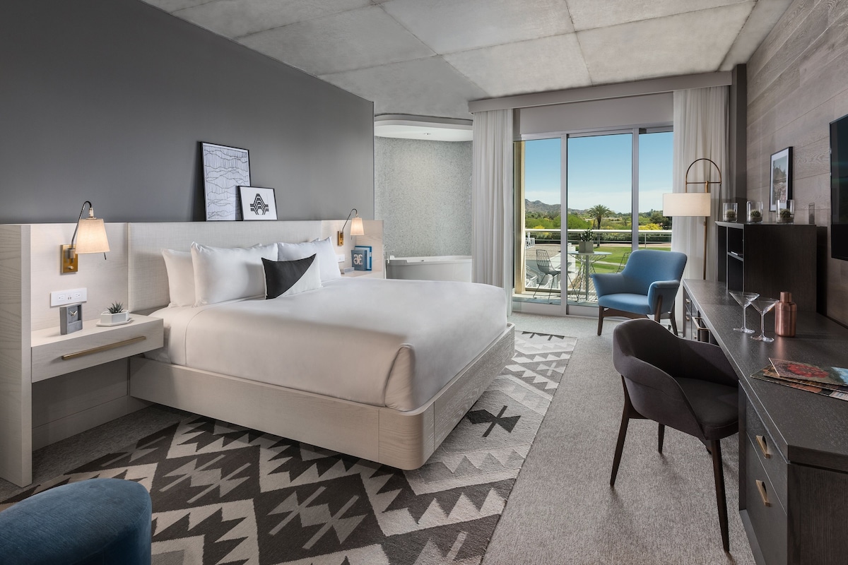 A modern hotel room with a large bed, a desk, a chair, and a balcony with a view.