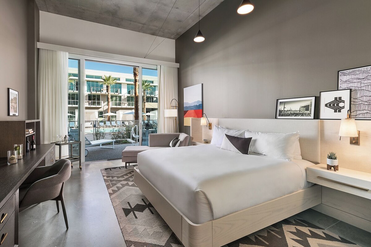 A modern hotel room with a large bed, a desk, and a view of a pool and building.