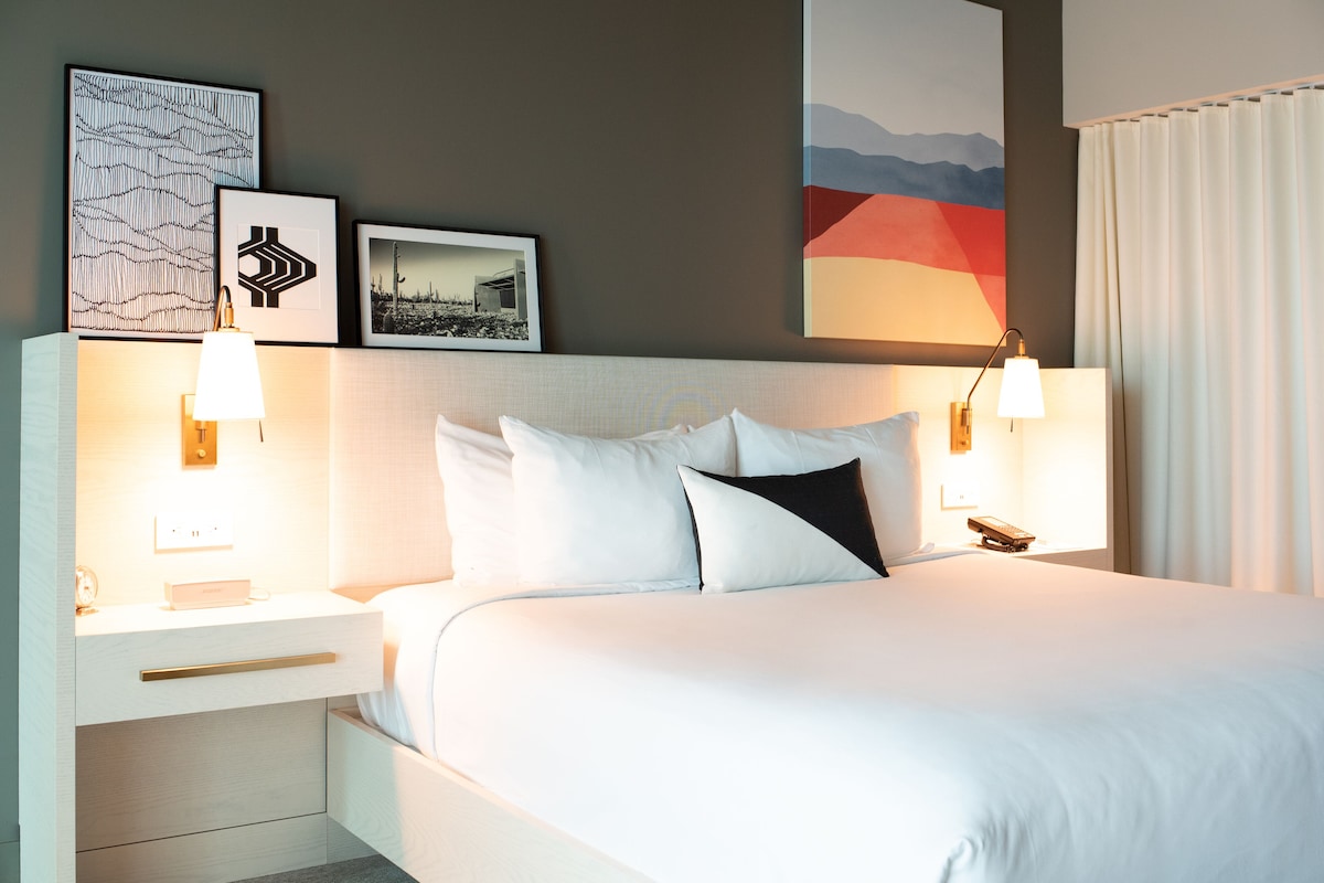 A neatly made bed with white bedding and black pillows, a bedside table with a lamp, and framed artwork on the wall.