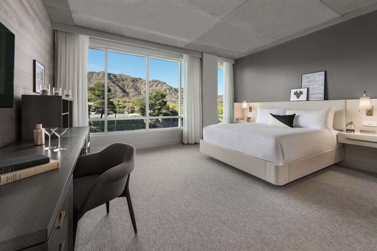 A modern hotel room with a large bed, a desk with a chair, a TV, and a view of mountains through the window.