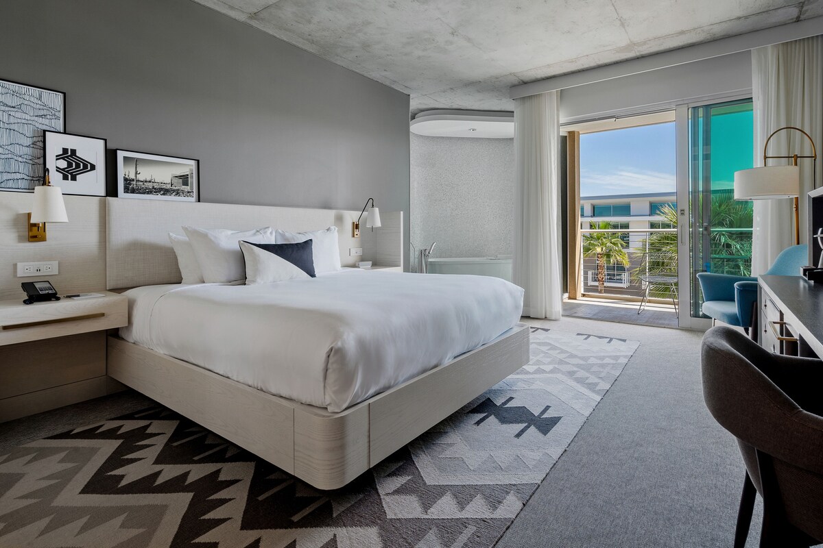 A modern hotel room with a large bed, a desk, a chair, and a balcony with a view of palm trees.