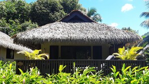 Una cabana amb teulada de palla i un porxo de fusta, envoltada de vegetació abundant i palmeres.