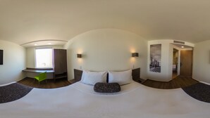 A panoramic view of a modern hotel room with a large bed, a desk, a chair, and a TV mounted on the wall. - One Guadalajara Expo (Guadalajara)