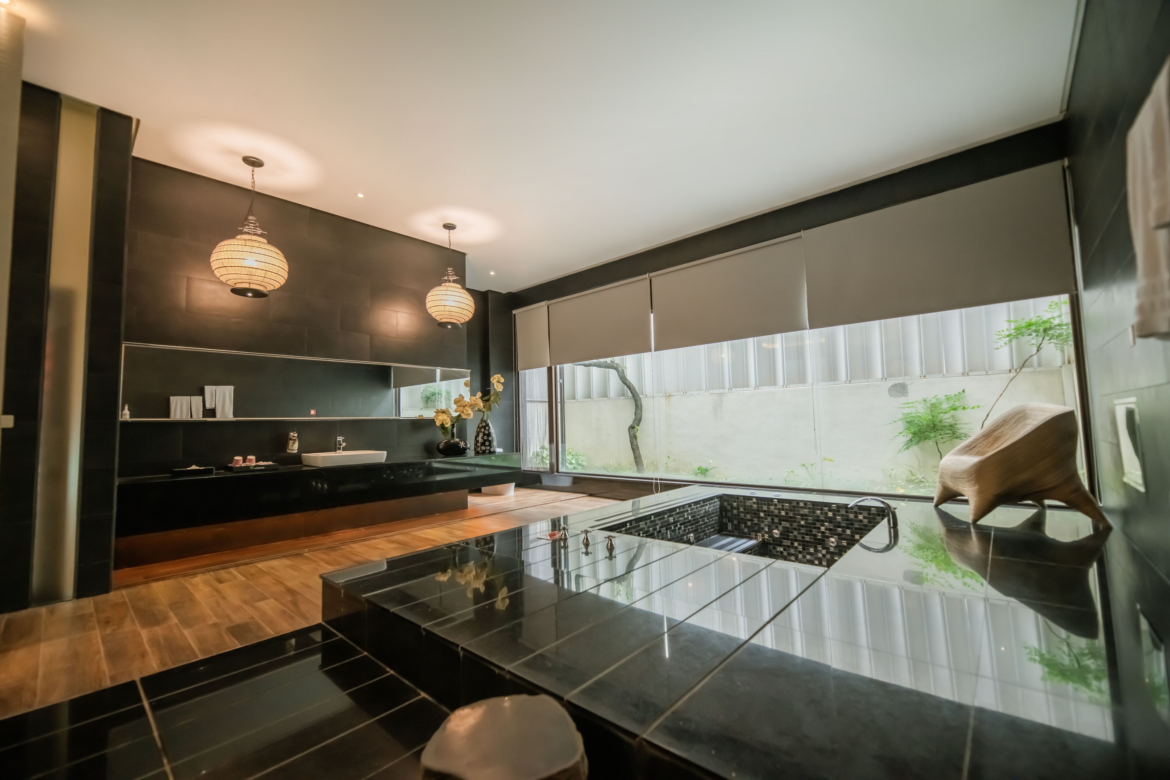 A modern bathroom with a large mirror, two pendant lights, a black sink, and a small seating area.