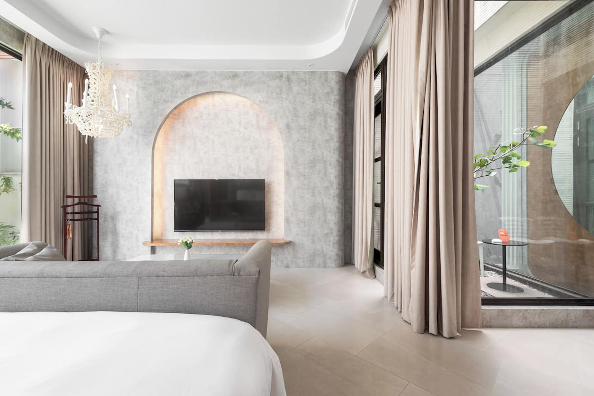 A modern bedroom with a large bed, a TV mounted on a stone wall, a small table with a vase, and a view of the outdoors through a glass window.