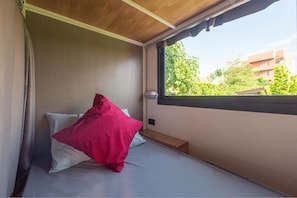 Una habitación pequeña con una cama individual, ventana, techo de madera y vista a espacios verdes.