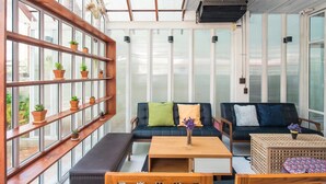 A modern living room with a glass ceiling, wooden shelves with potted plants, a black sofa with cushions, a wooden coffee table, and a patterned ottoman. - Haus Hostel (Chiang Mai)
