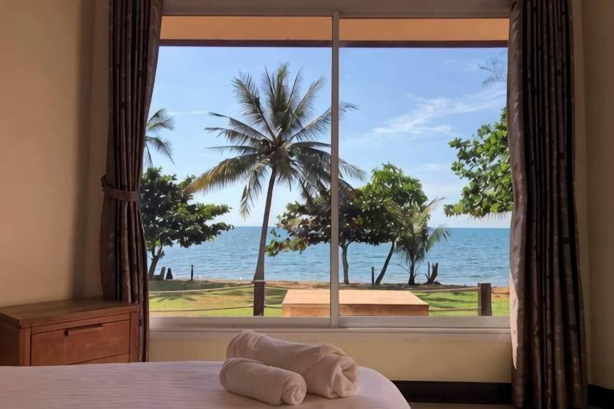 Un dormitorio con un ventanal grande, una cama y vistas al océano y palmeras.