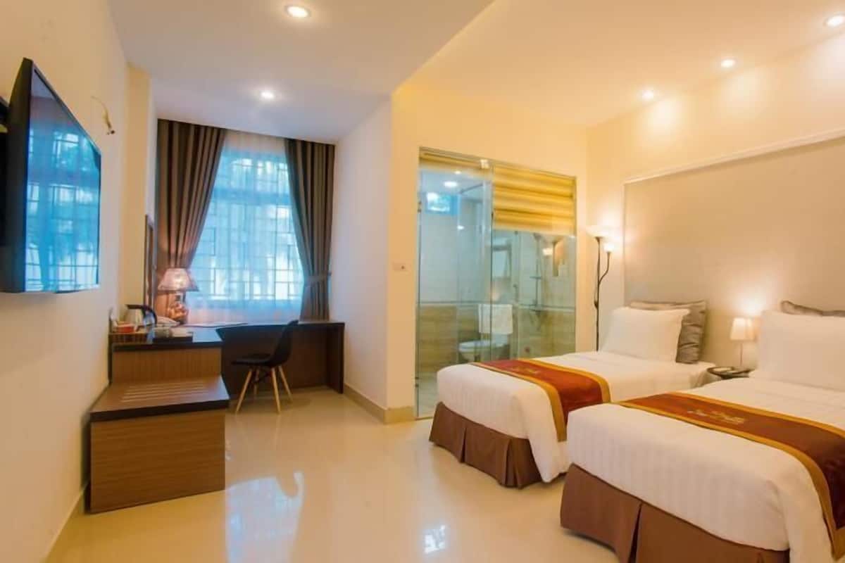 Habitación de hotel con dos camas, un escritorio, una silla, un televisor y un baño visible a través de una puerta de cristal.