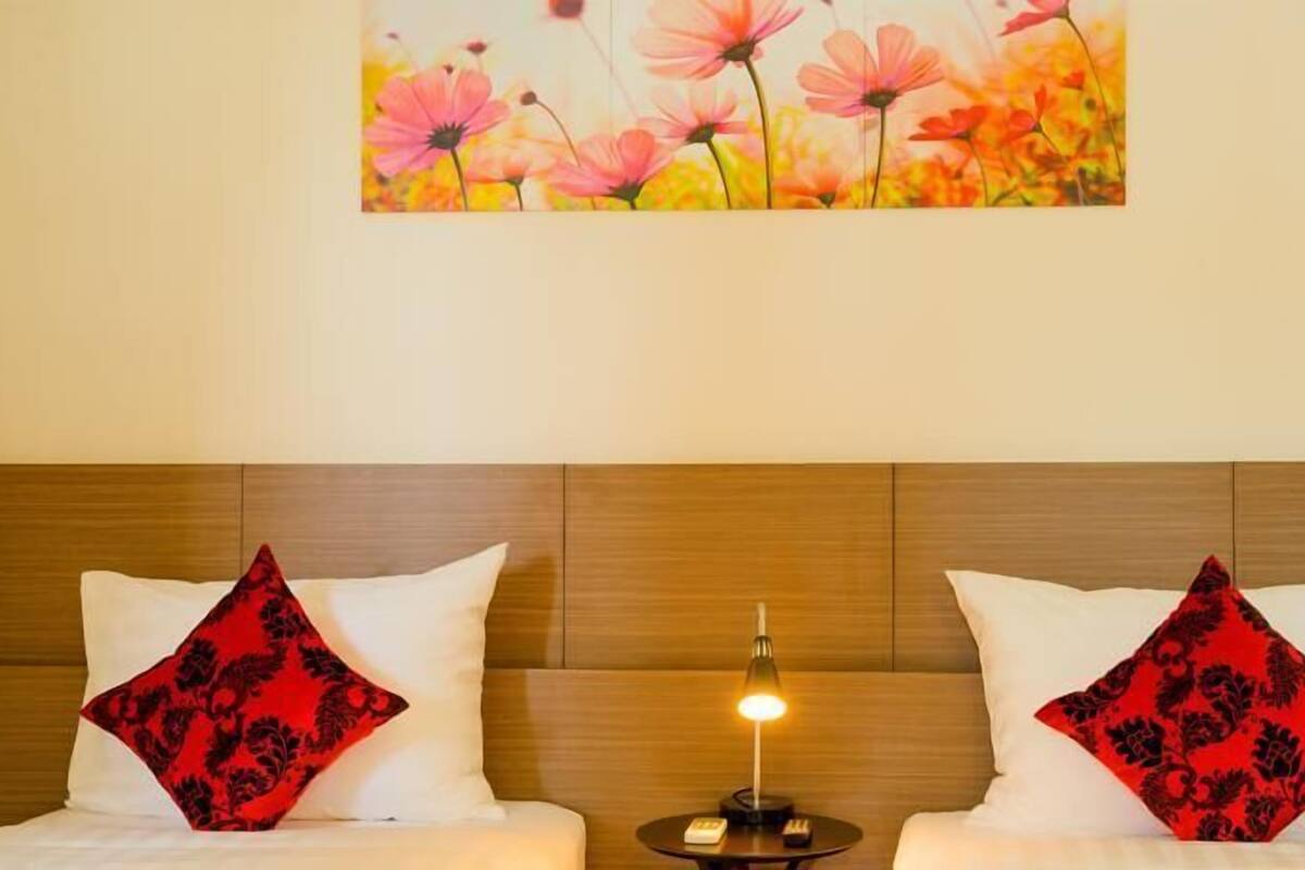Habitación de hotel con cabecera de madera, dos almohadas blancas con motivos florales rojos, una mesita de noche con lámpara y un cuadro de flores rosas y amarillas en la pared.