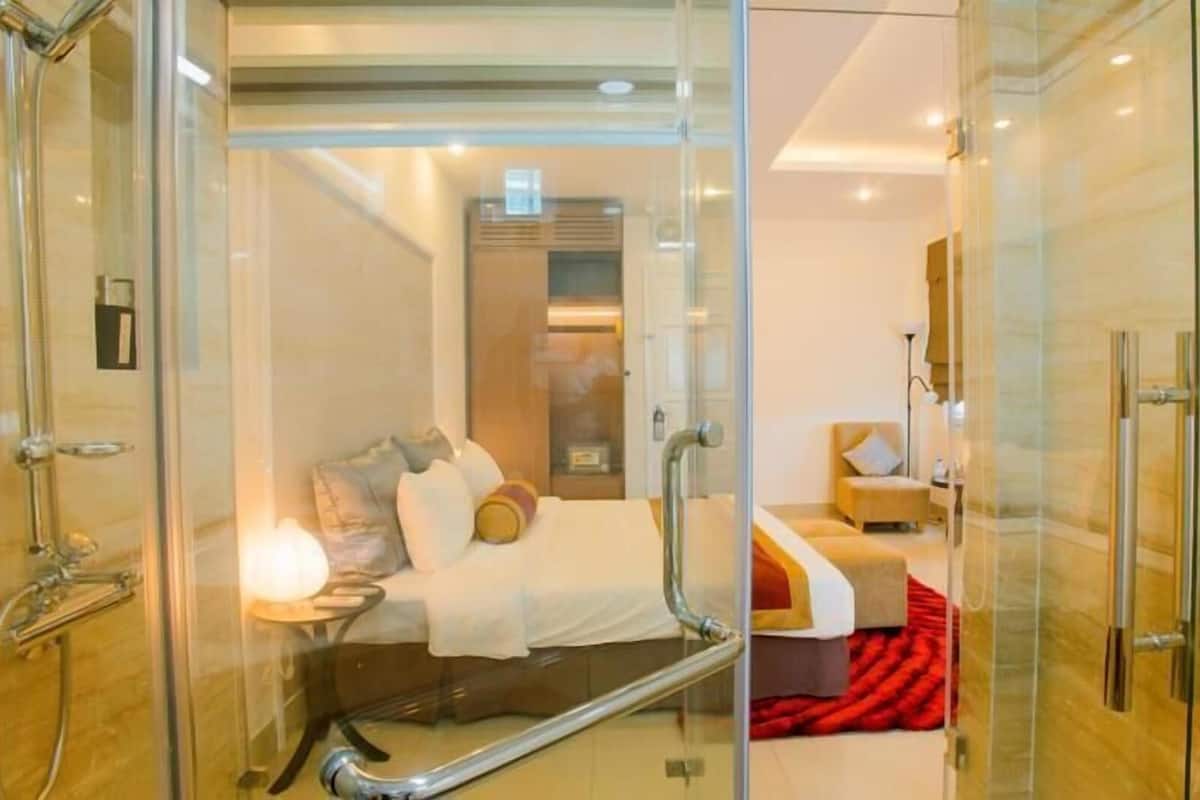 Una habitación de hotel moderna con cabina de ducha de vidrio, cama con almohadas y manta, y una pequeña zona de estar con una silla y una mesa.