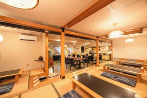 Une auberge japonaise traditionnelle avec des tatamis au sol, des meubles en bois et un coin cuisine à l’arrière-plan.