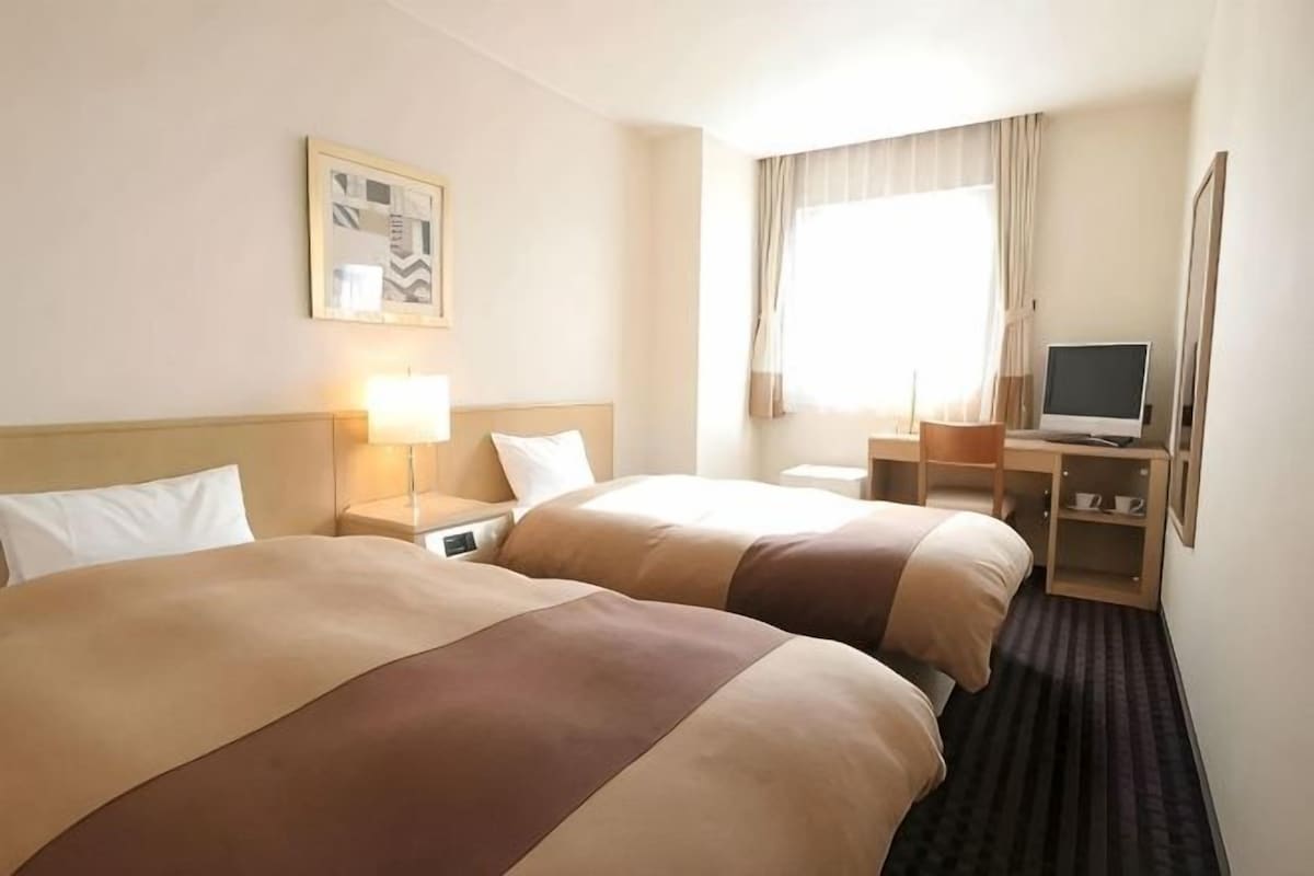 Une chambre d’hôtel avec deux lits, un bureau avec un ordinateur, une fenêtre avec des rideaux et un téléviseur fixé au mur.