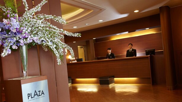Un espace d’accueil d’hôtel avec un comptoir de réception en bois, deux employés et une composition florale.