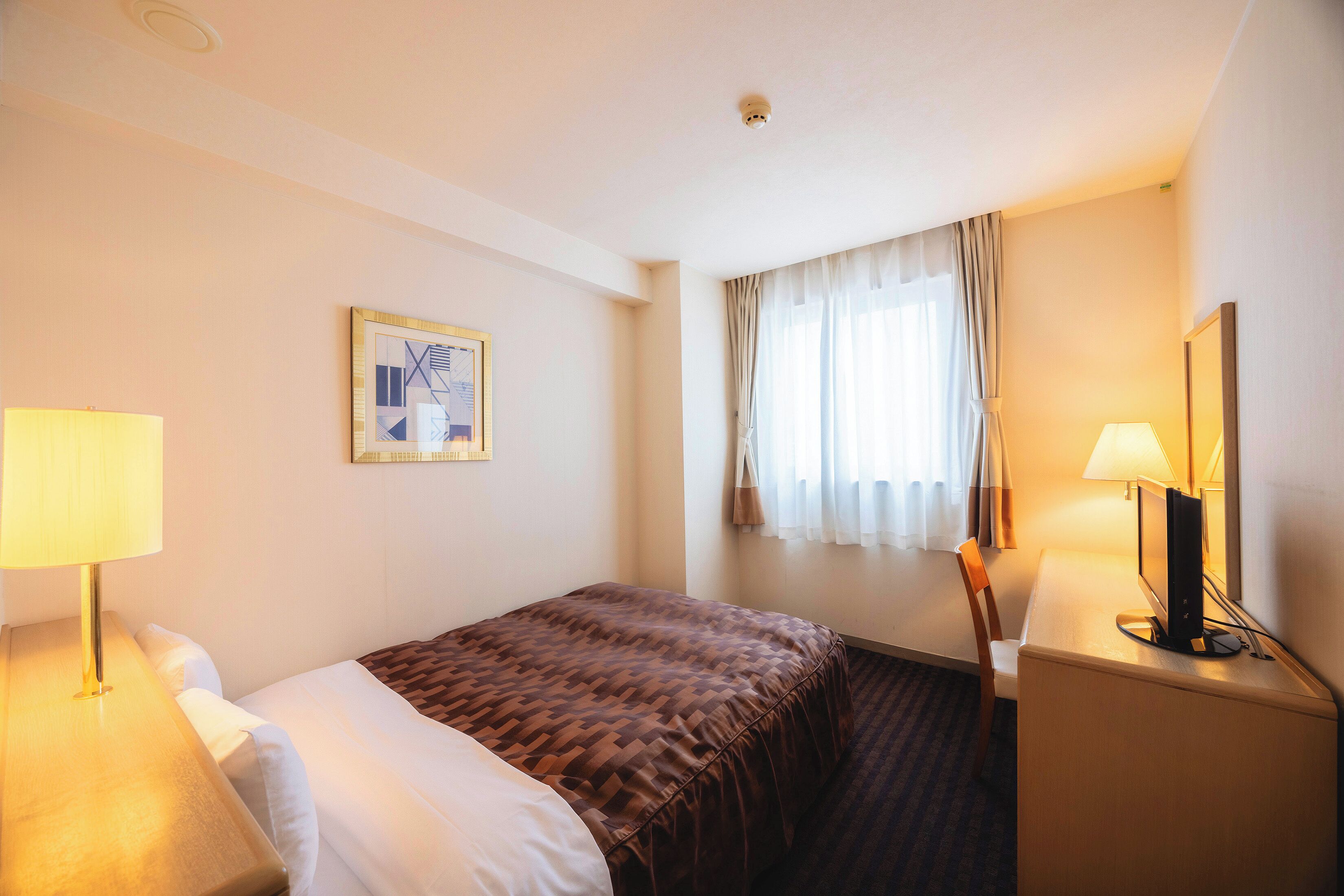 Quarto de hotel com cama, escrivaninha com computador, luminária e janela com cortinas.